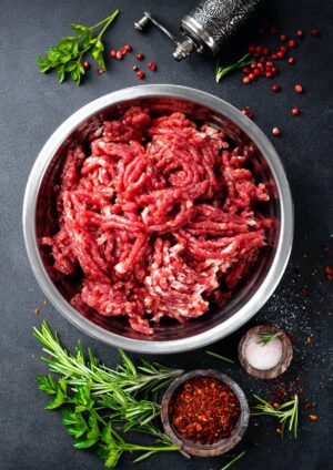 A bowl of raw ground meat, likely ground sirloin or ground chuck, surrounded by parsley, rosemary, salt, and pepper on a dark surface.