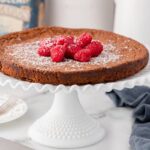 A flourless chocolate cake tart is displayed on a white cake stand, topped with powdered sugar and fresh raspberries. A fork, knife, and blue cloth are placed nearby.