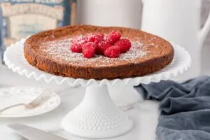 A flourless chocolate cake tart is displayed on a white cake stand, topped with powdered sugar and fresh raspberries. A fork, knife, and blue cloth are placed nearby.