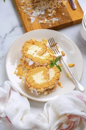 A dessert dish featuring two slices of rolled cake filled with cream, garnished with sliced almonds, mint leaves, and accompanied by a fork, placed on a white cloth on a marble surface.