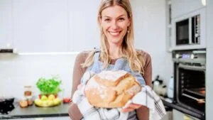 A smiling woman wearing an apron holds a freshly baked loaf of sourdough bread in a kitchen.