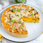 A whole savory sweet potato pie with one slice missing, topped with herbs and grated cheese, on a white plate.